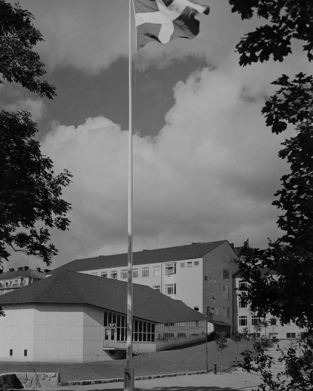 Svartvitt fotografi av en flaggstång med flagga framför en skolbyggnad