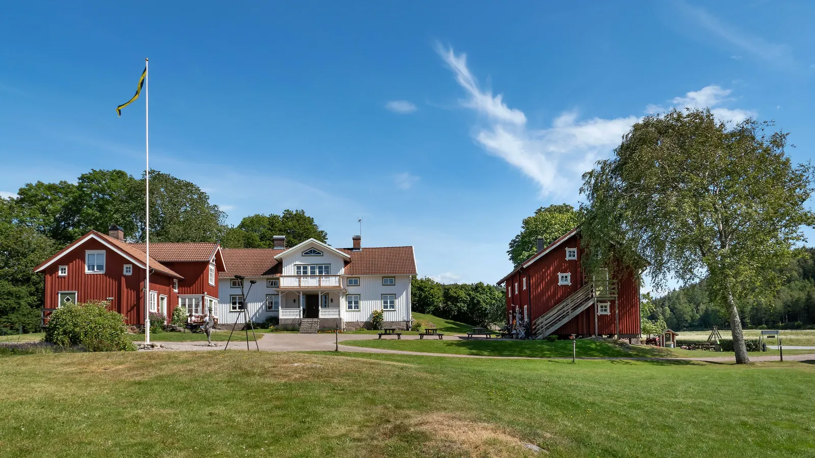 Fristående flaggstång med svensk flagga på en gårdsplan framför röda och vita gårdshus